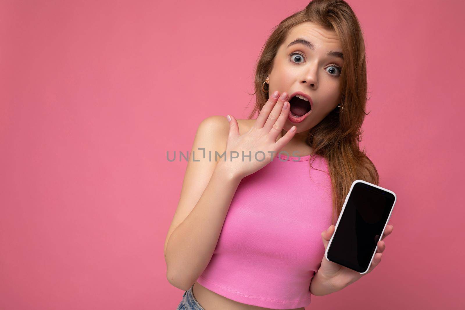 Photo of amazed beautiful joyful young blonde woman wearing pink top poising isolated on pink background with empty space holding in hand and showing mobile phone with empty display for mockup looking at camera.