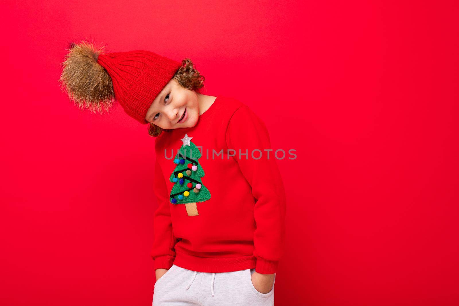 cute blond boy in warm hat and christmas sweater on red background with smile on his face.