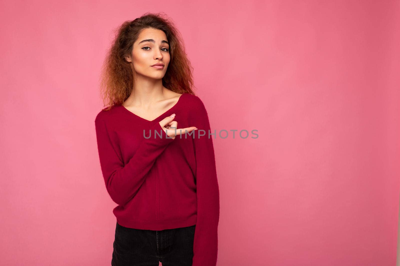 Young beautiful brunette wavy woman with sincere emotions wearing casual pink jersey isolated over pink background with copy space and pointing at free space for mockup.