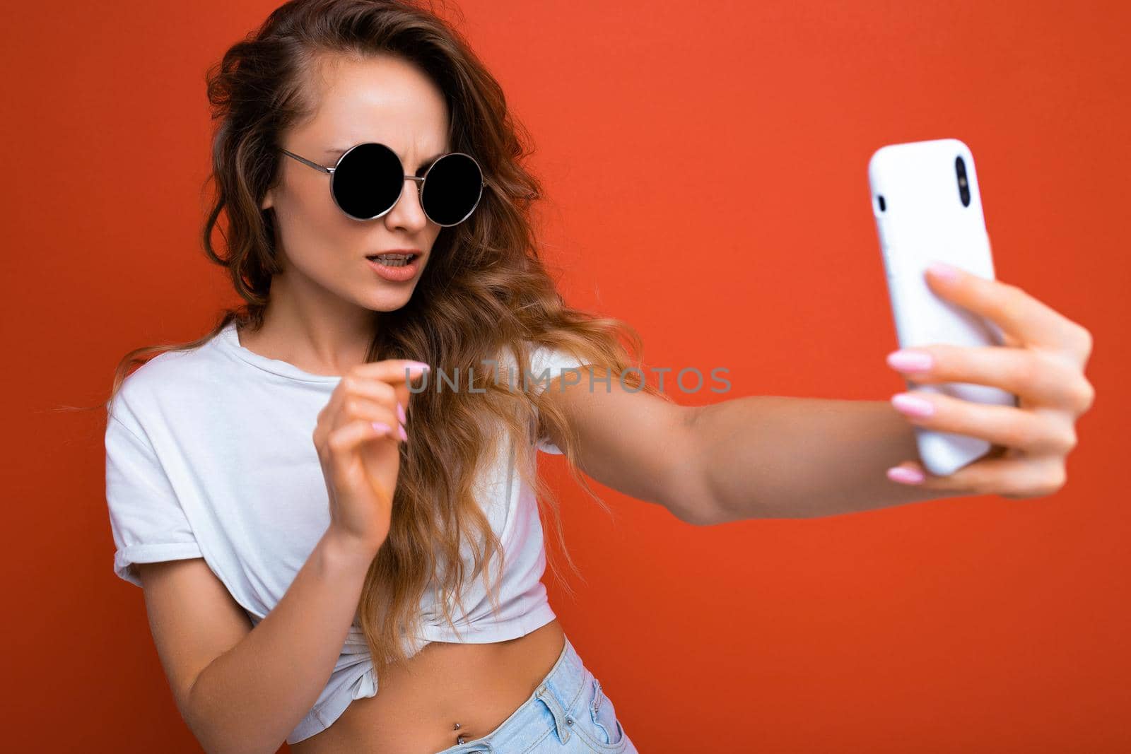 Amazing beautiful young blonde woman holding mobile phone taking selfie photo using smartphone camera wearing everyday stylish outfit isolated over colorful wall background looking at device screen.