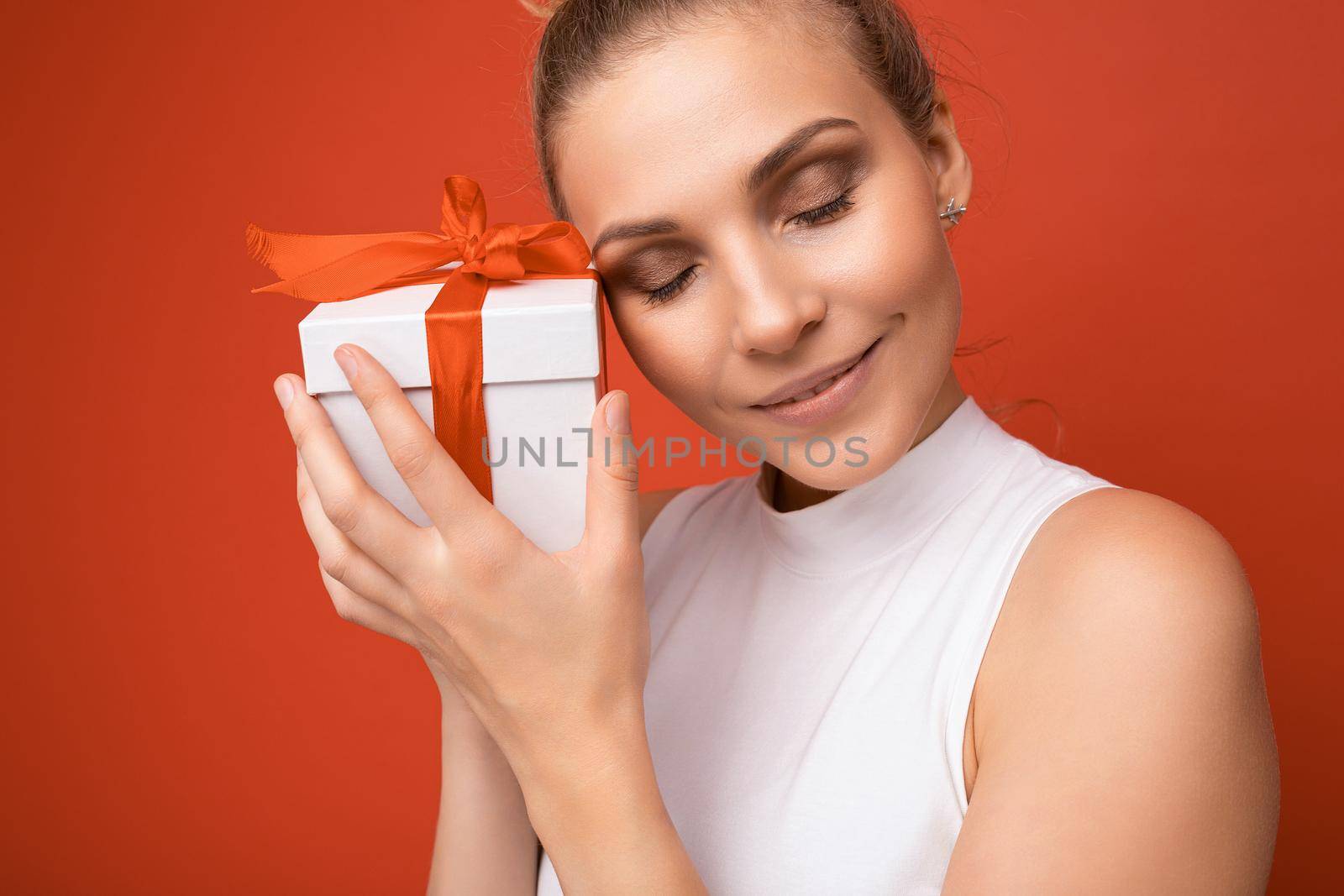 Portrait photo shot of attractive happy young blonde woman isolated over red background wall wearing white top holding gift box and enjoyig with close eyes.