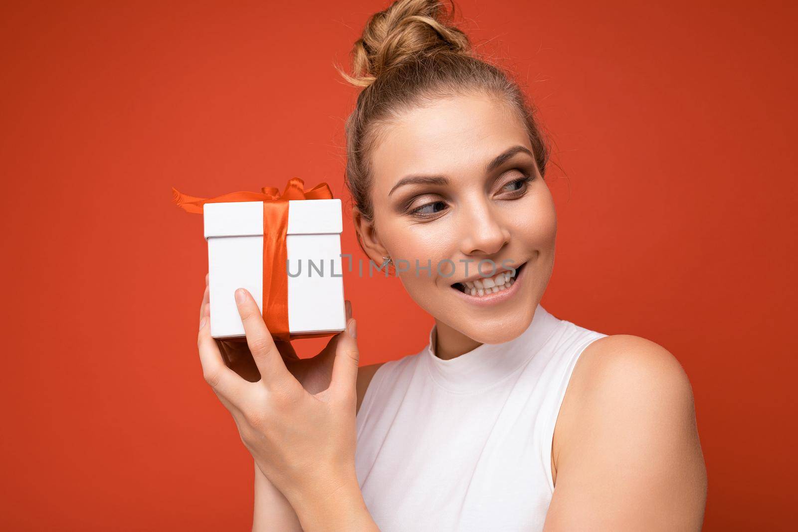 Portrait photo shot of attractive happy emotional young blonde woman isolated over red background wall wearing white top holding gift box and looking to the side.