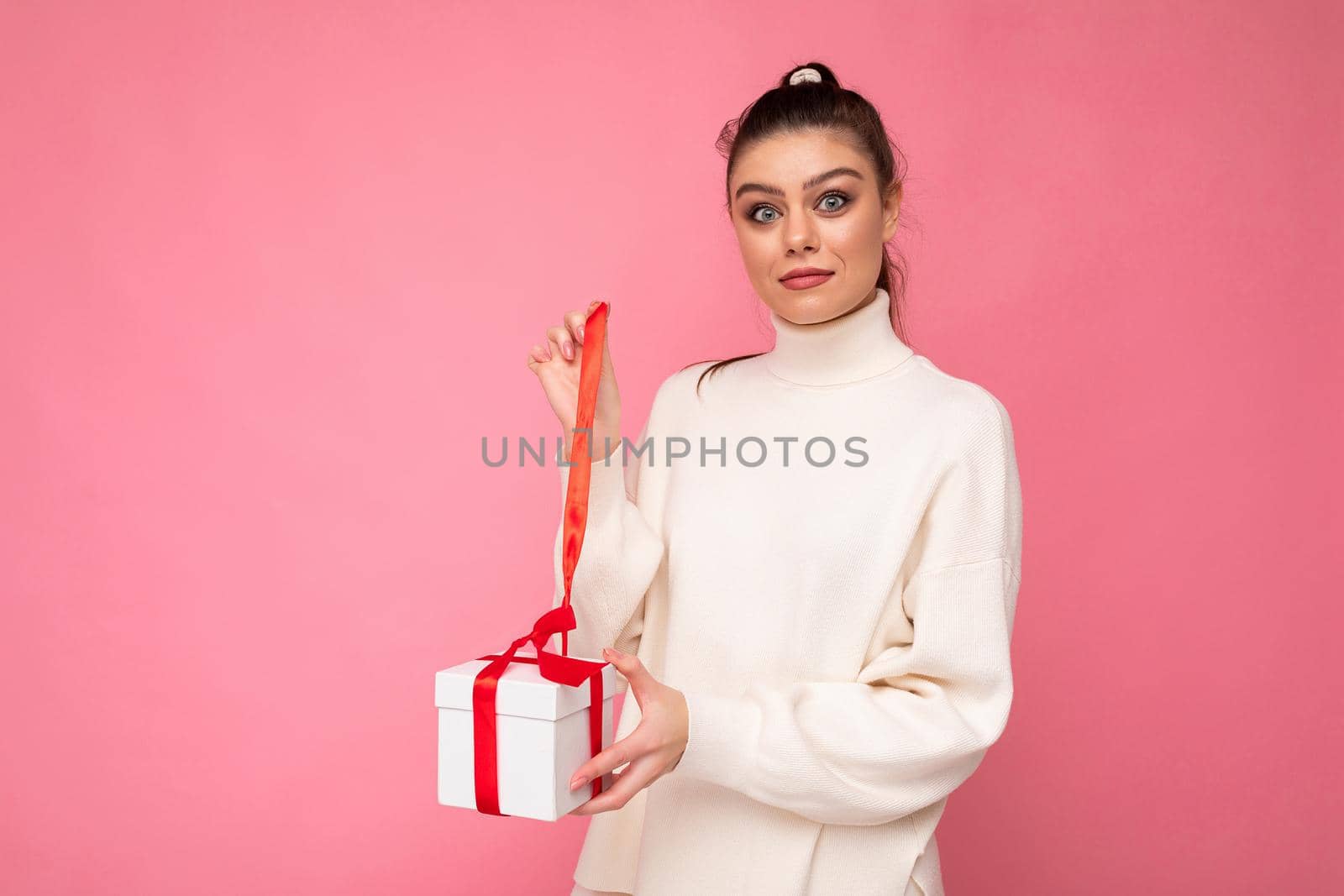 Attractive positive surprised young brunette woman isolated over pink background wall wearing white sweater holding gift box and unboxing present looking at camera.