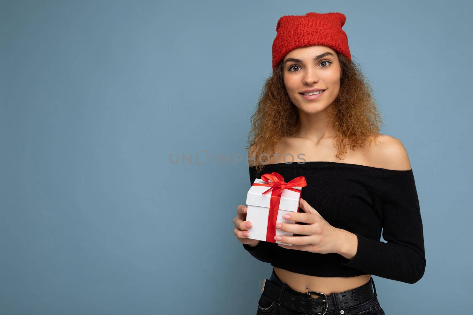 Photo shot of beautiful positive surprised young dark blonde curly woman isolated over blue background wall wearing black crop top and red hat holding gift box looking at camera. Empty space