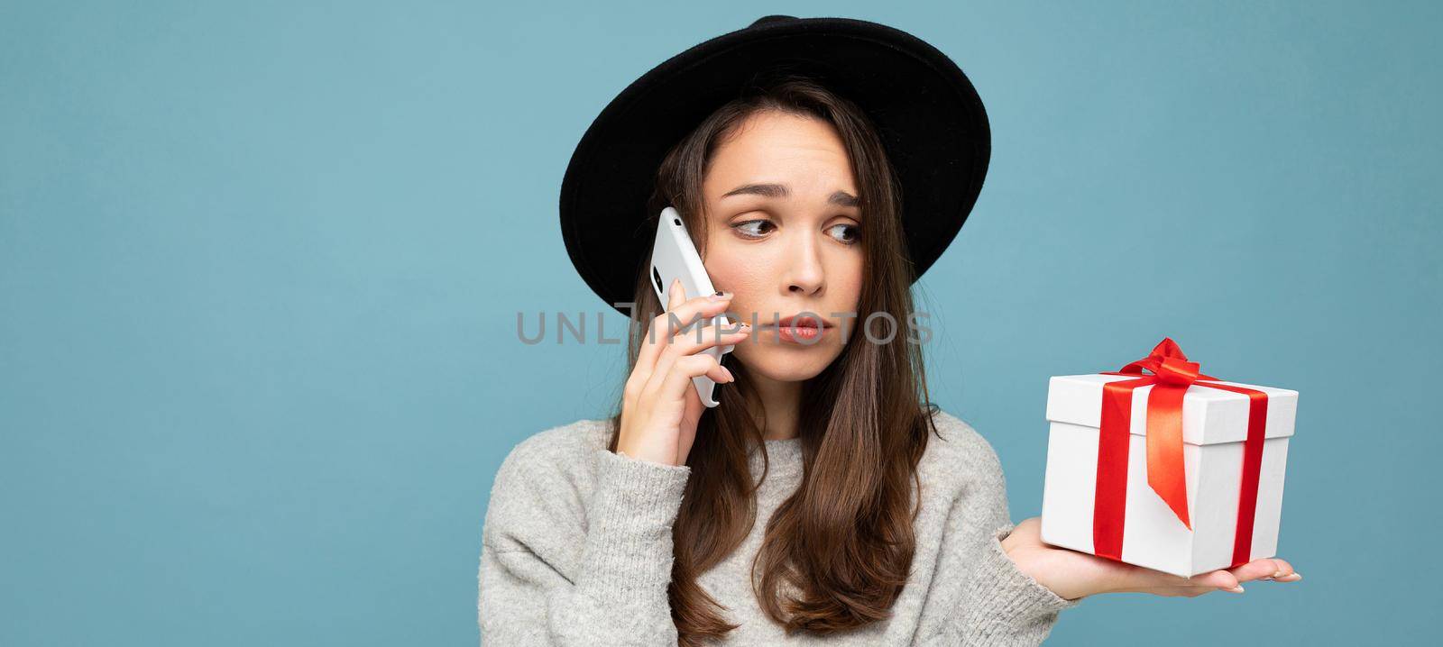 Photo of beautiful upset young brunette woman isolated over blue background wall wearing black hat and grey sweater holding gift box talking on mobile phone and looking to the side by TRMK