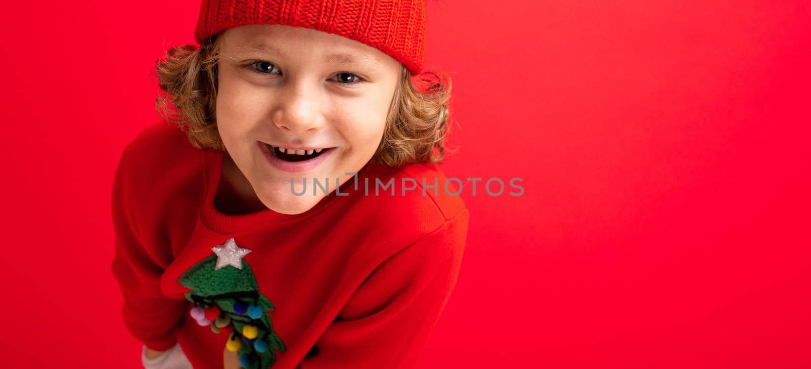 cool blond kid in warm hat and sweater with christmas tree on red background fooling around, christmas concept by TRMK