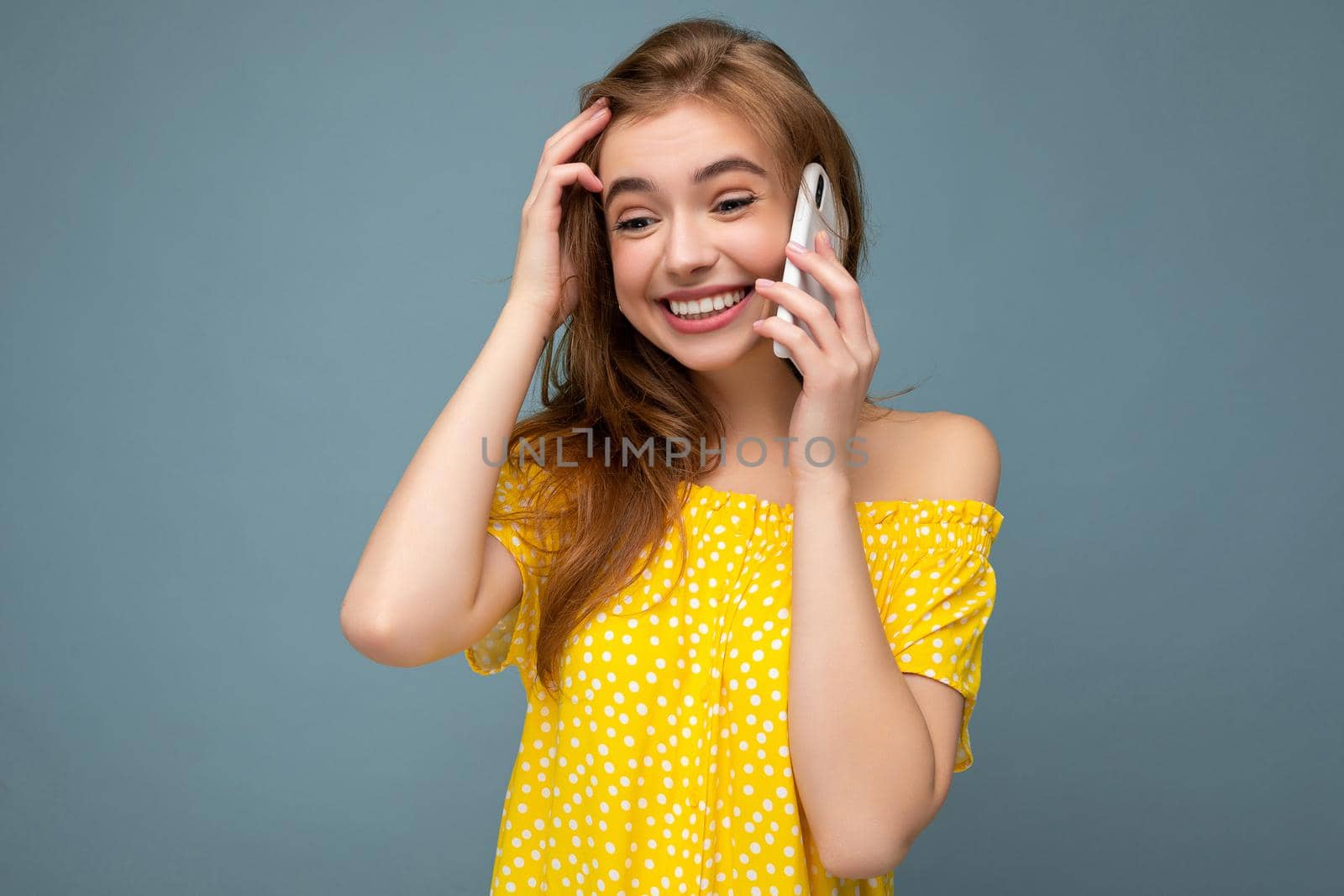 Attractive positive smiling young blonde woman wearing stylish yellow summer dress standing isolated over blue background holding and talking on mobile phone looking to the side by TRMK