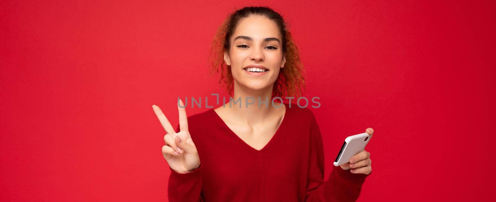Photo of happy smiling woman with gathered curly hair wearing dark red sweater isolated over red background holding smartphone having fun and showing peace gesture looking at camera.