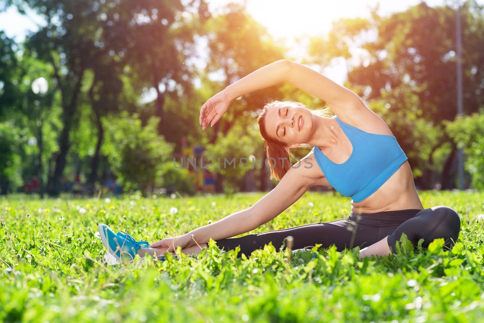 Attractive girl in sportswear doing yoga in park. Young woman sitting in yoga pose on green grass. Sport training outdoor at sunny summer day. Morning stretching exercises and healthy lifestyle.