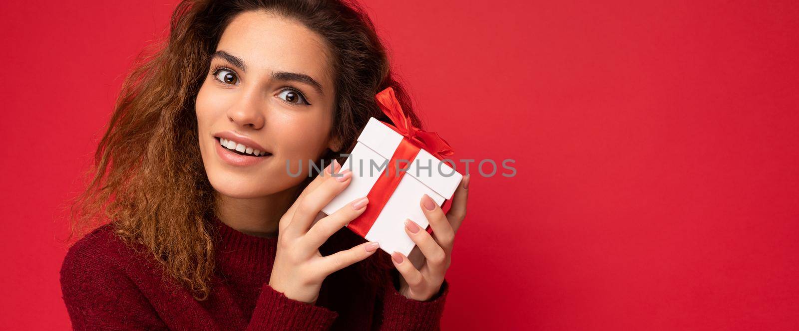 Closeup photo of attractive positive smiling young brunette curly woman isolated over red background wall wearing red sweater holding gift box looking at camera and dreaming. Copy space