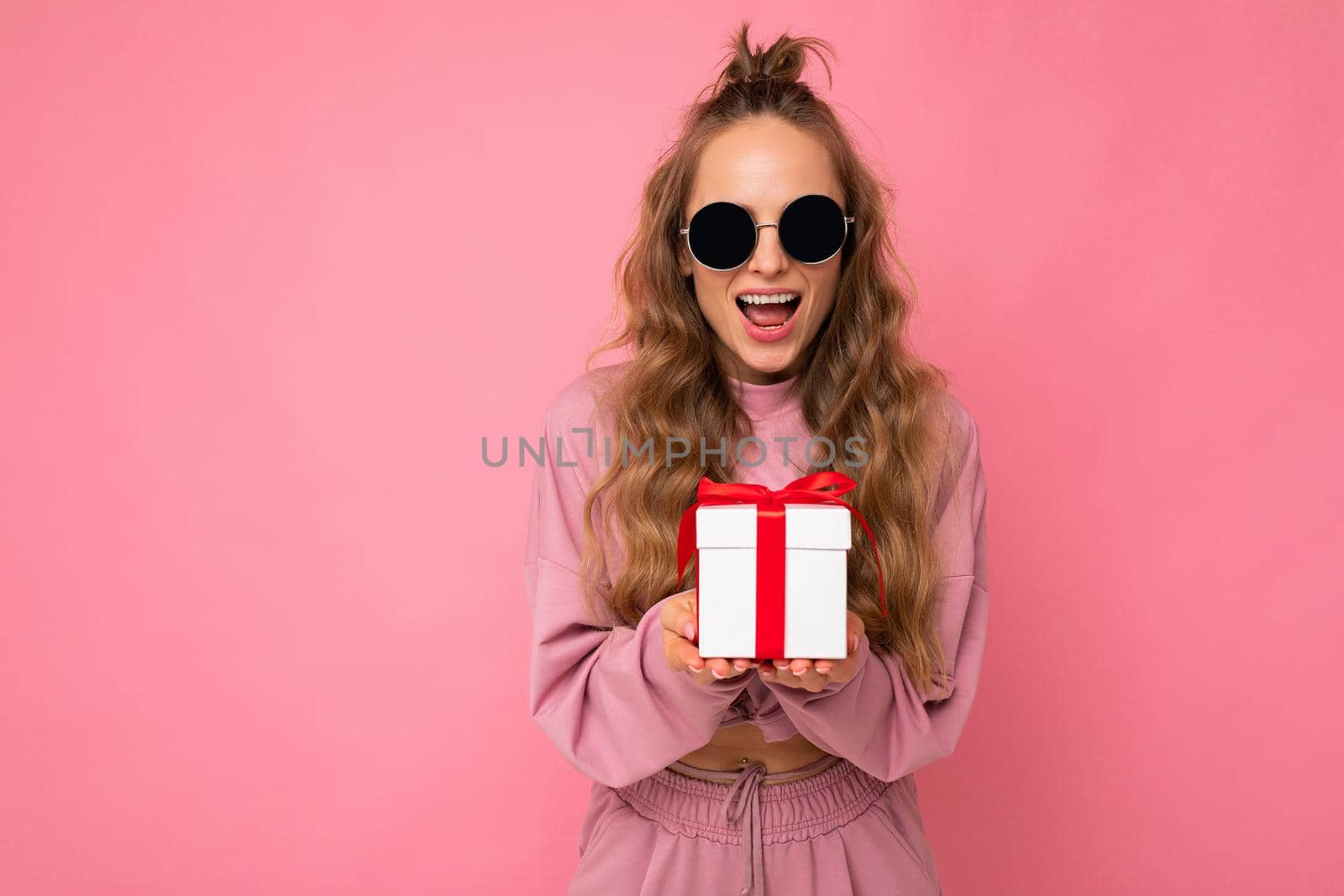 Beautiful happy young brunette woman isolated over colourful background wall wearing stylish casual clothes holding gift box and looking at camera. Copy space