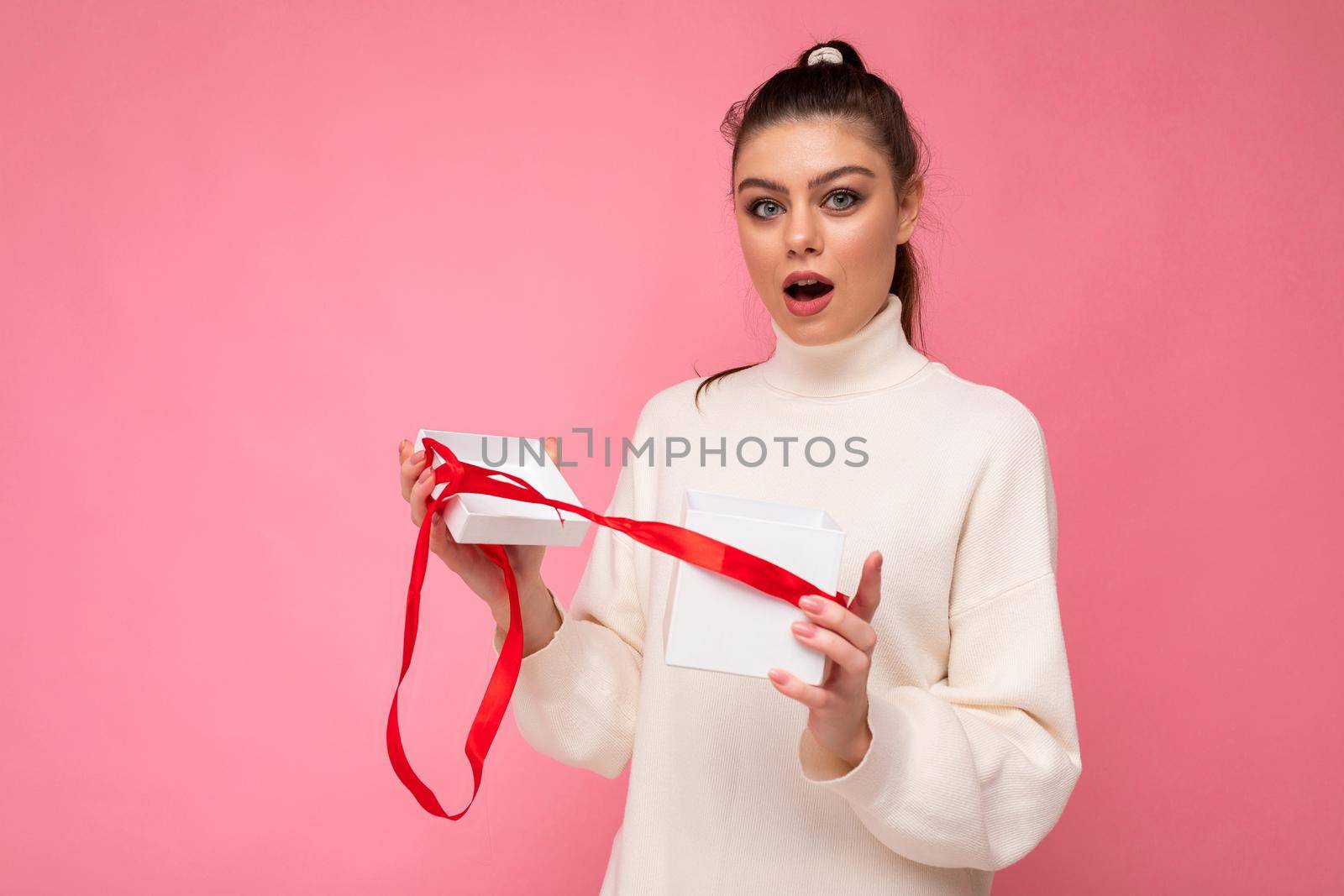 Fascinating amazed astonished adult brunette woman isolated over pink background wall wearing white sweater holding gift box and unpacking surprise looking at camera.
