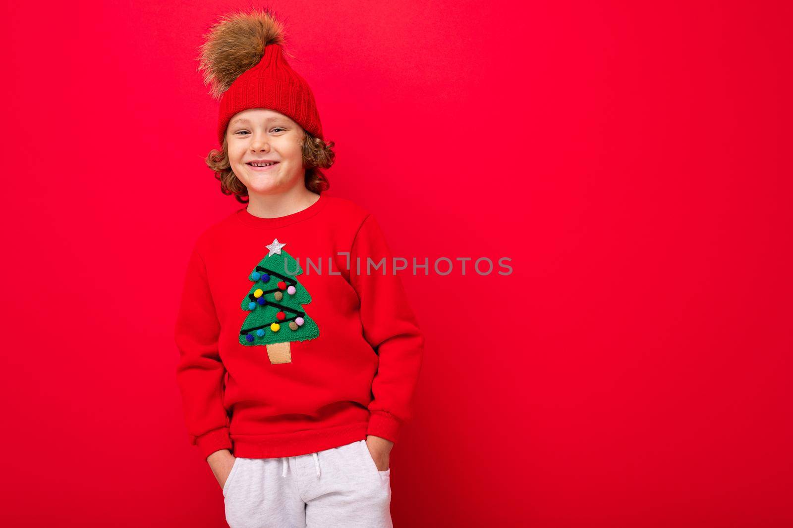 cool blond kid in warm hat and sweater with christmas tree on red background fooling around, christmas concept.