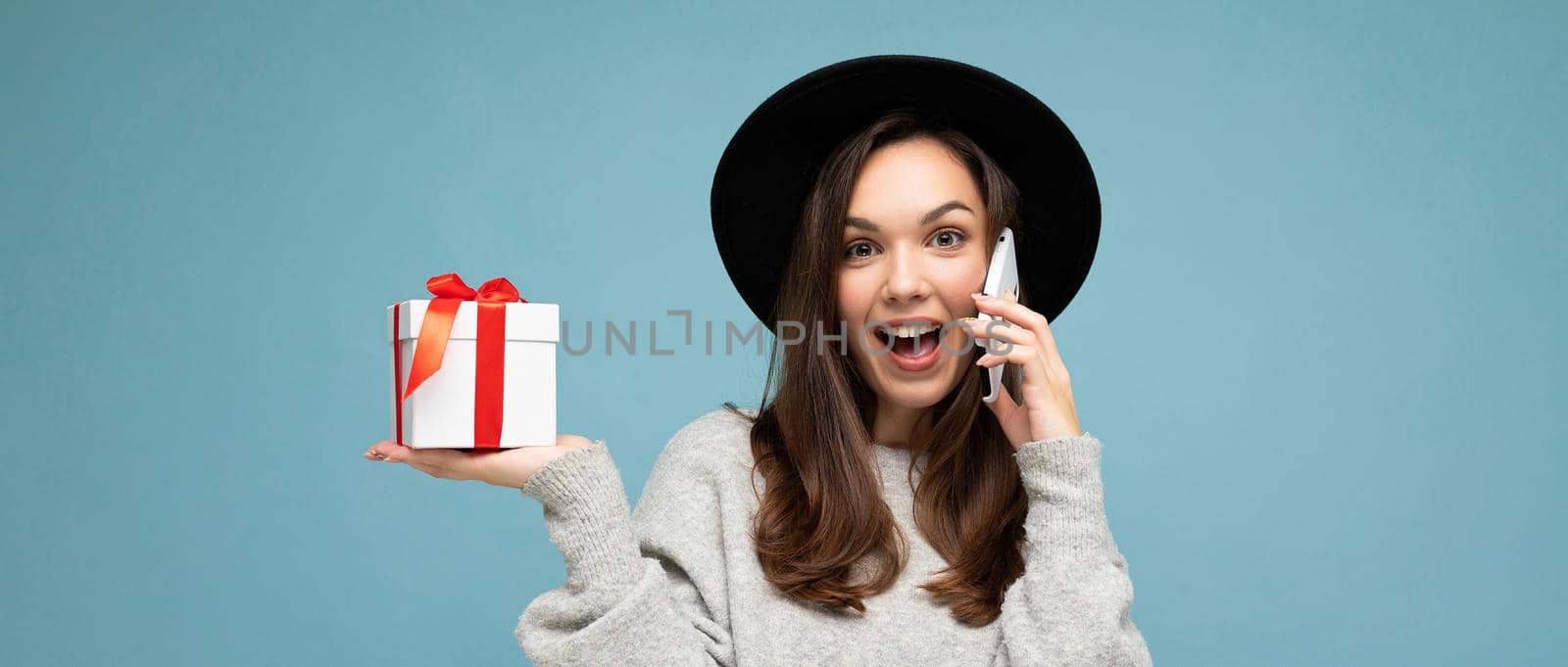 Panoramic shot of attractive happy positive funny young brunette woman isolated over blue background wall wearing black hat and grey sweater holding gift box talking on mobile phone and looking at camera by TRMK