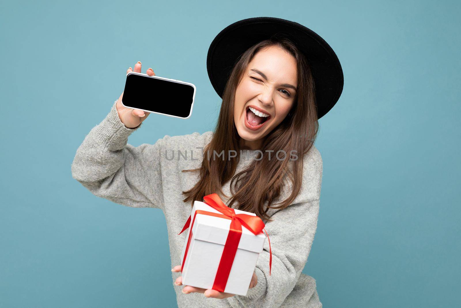 Attractive happy joyful young brunette woman isolated over blue background wall wearing fashionable black hat and casual grey sweater holding gift box showing mobile phone screen display for mockup and looking at camera and winking.