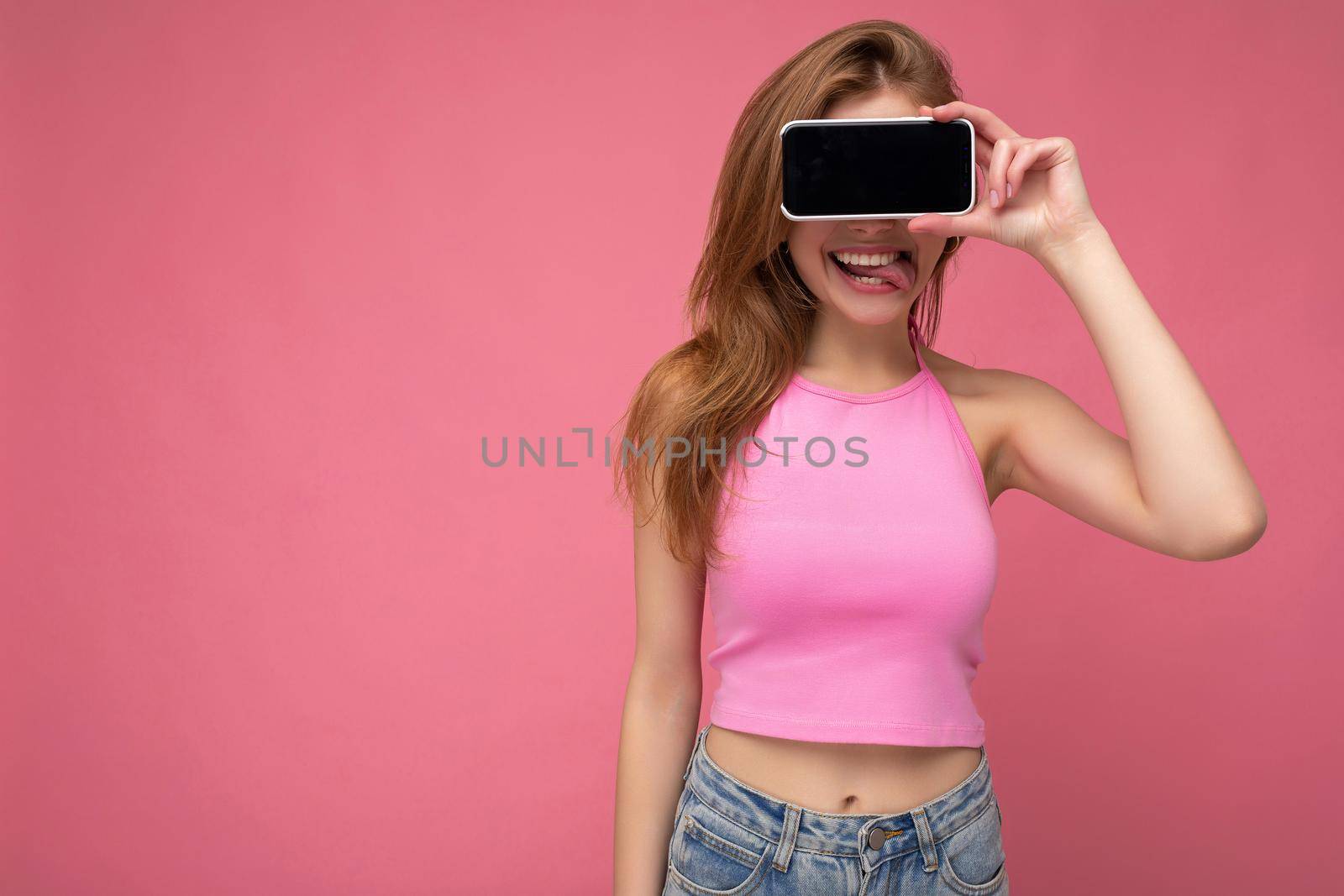 Photo of beautiful positive young blonde woman wearing pink top poising isolated on pink background with empty space holding in hand and showing mobile phone with empty display for mockup and shoeing tongue.