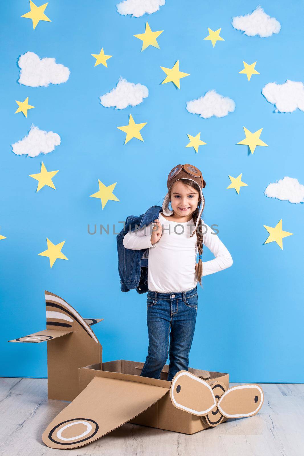 Little child girl in a pilot's costume is playing and dreaming of flying over the clouds. Portrait of funny kid on a background of bright blue wall with yellow stars and white clouds