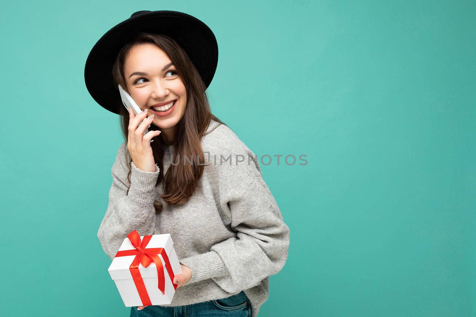 Photo of beautiful happy positive joyful young brunette woman isolated over blue background wall wearing black hat and grey sweater holding gift box talking on mobile phone and looking to the side. Copy space, mockup