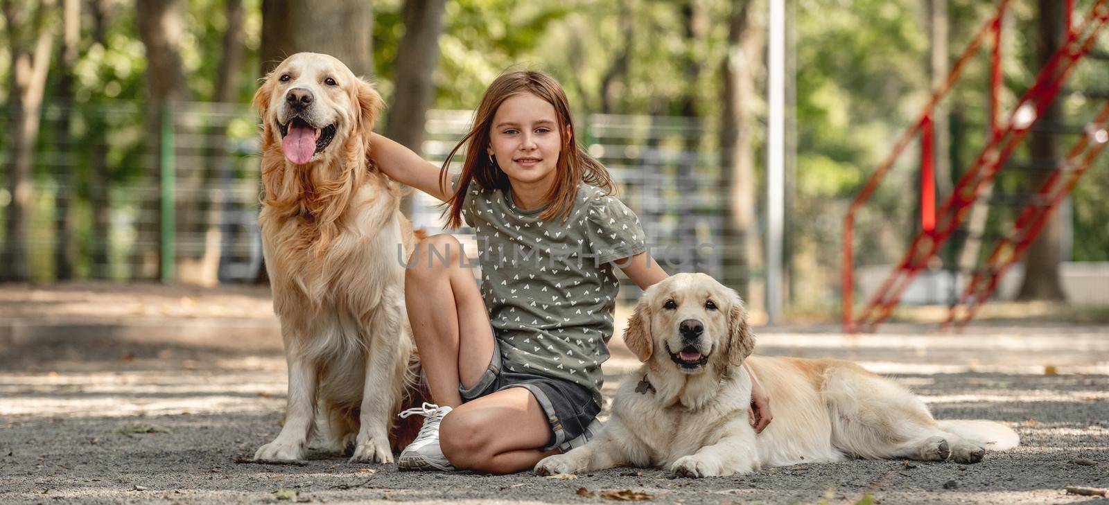 Preteen girl with golden retriever dogs sitting in the park. Cute child with doggy pets posing outdoors