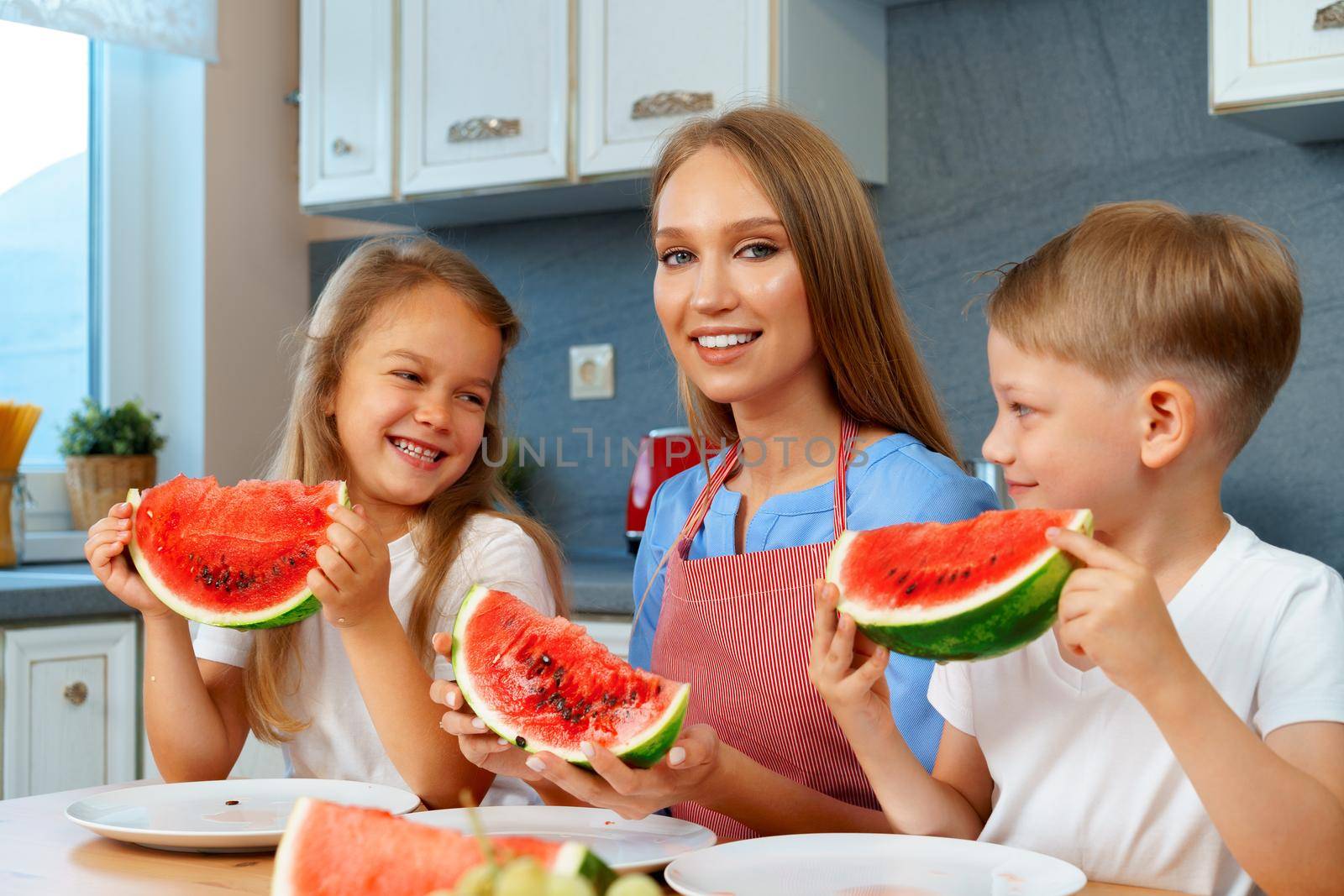 Sweet family, mother and her kids eating watermelon in their kitchen having fun by Fabrikasimf