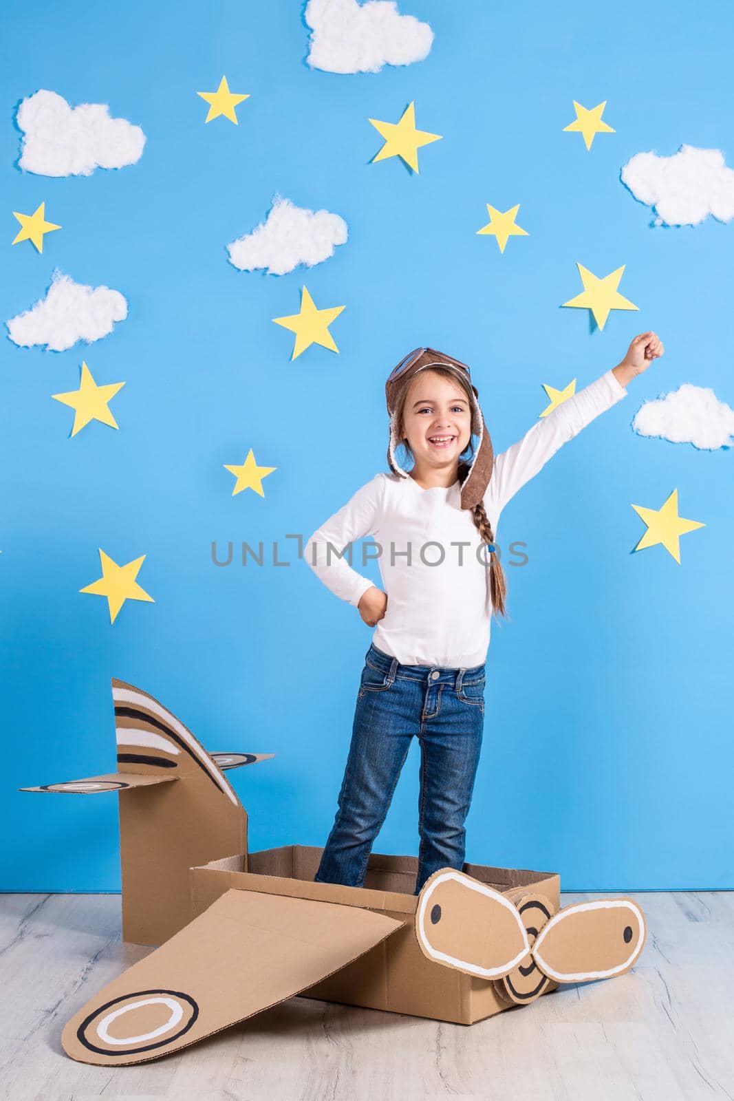 Little dreamer girl playing with a cardboard airplane at the studio with blue sky and white clouds background. Childhood. Fantasy, imagination.