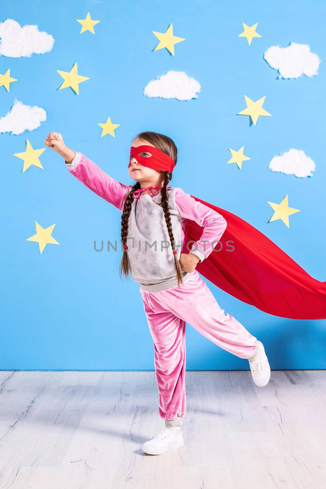 Six year blonde girl dressed like superhero having fun at home. Kid on the background of bright blue wall with white clouds and yellow stars. Girl power concept.