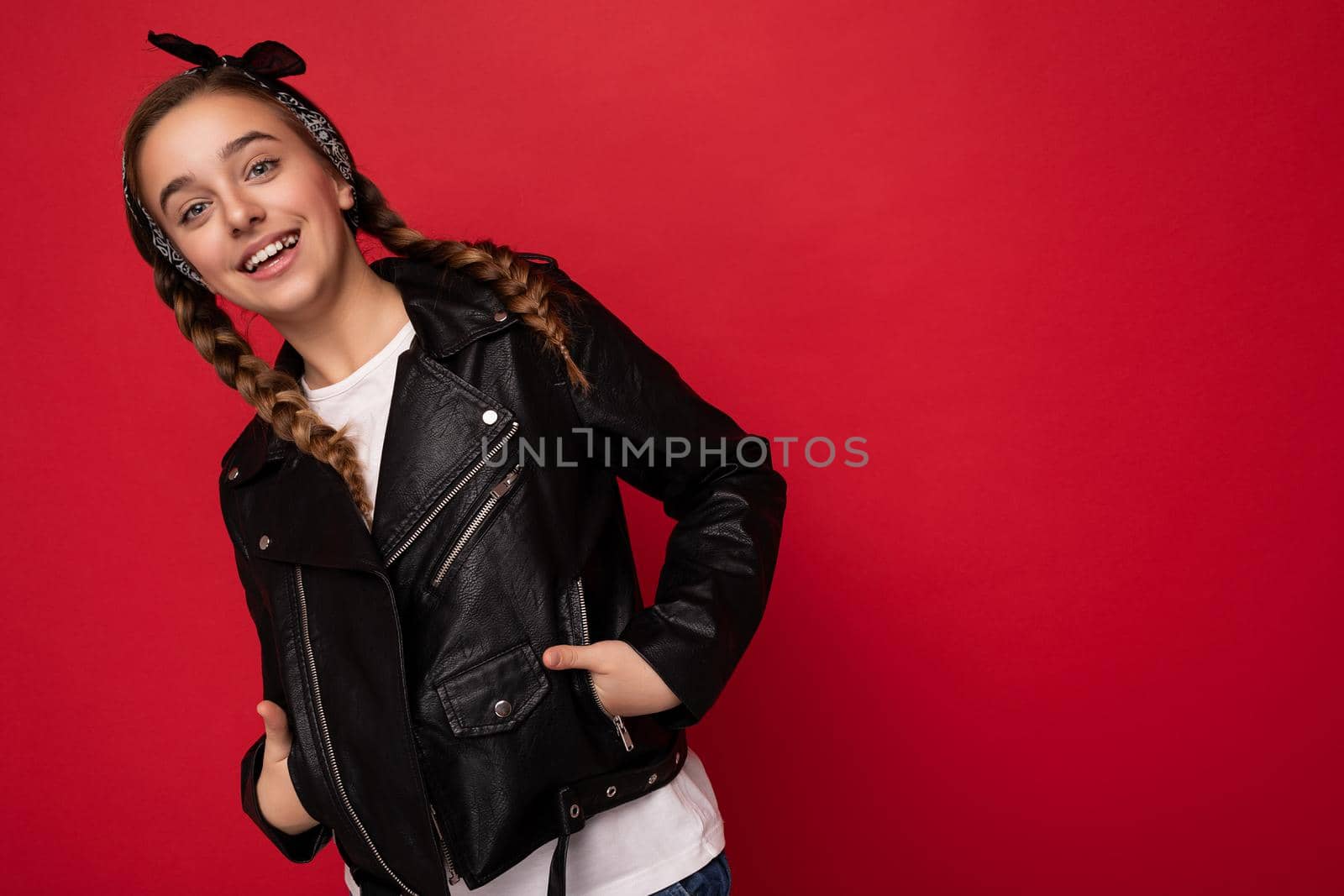 Attractive cool positive smiling brunetfemale teenager with pigtails wearing stylish black leather jacket and white t-shirt isolated over red background wall looking at camera. Copy space