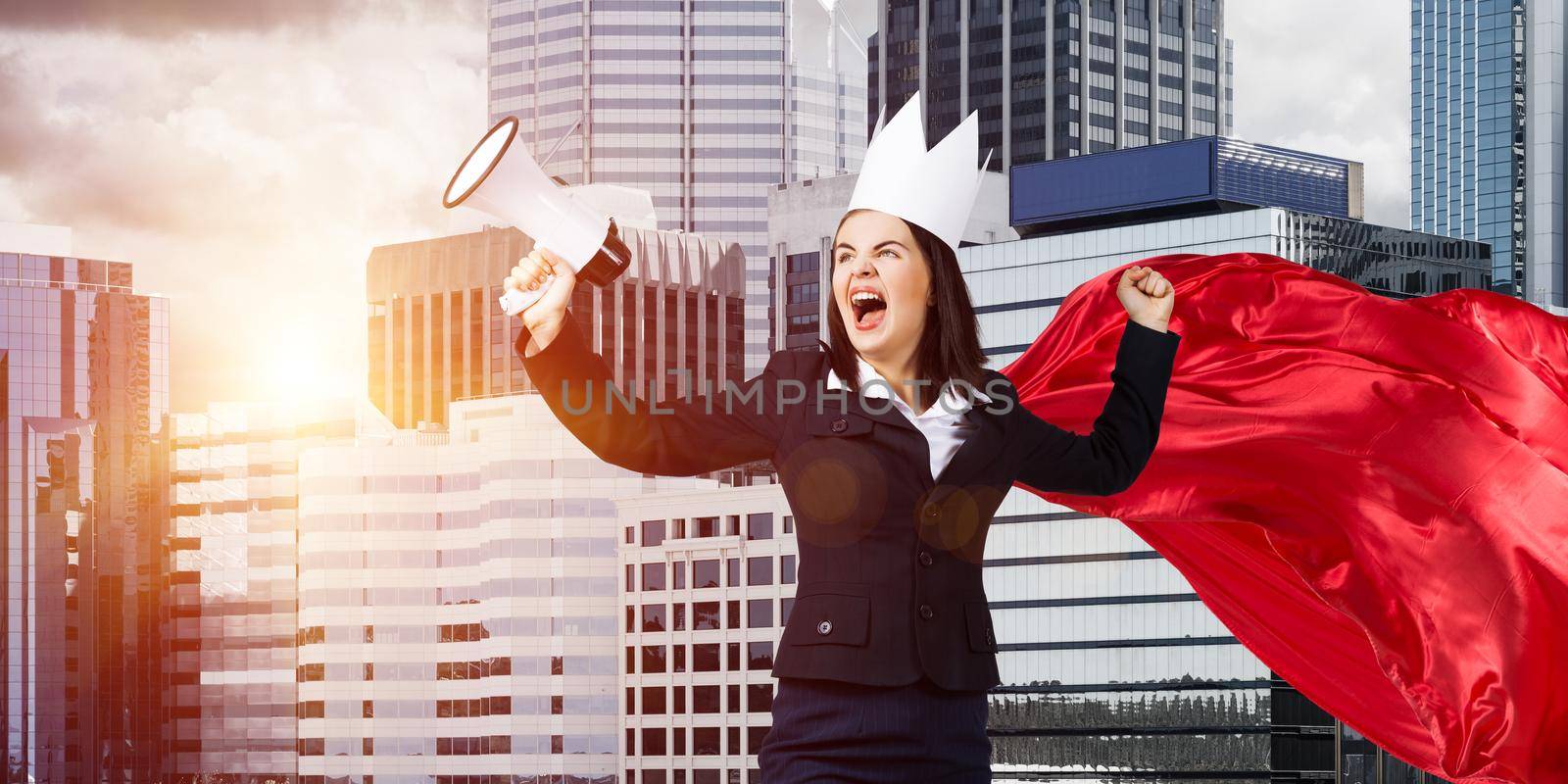 Young confident businesswoman wearing red cape against modern city background screaming in megaphone