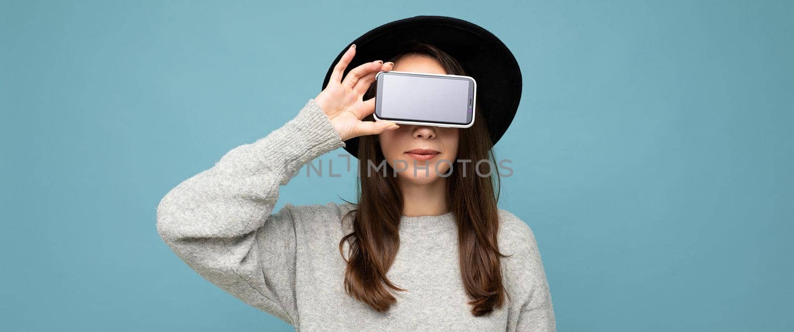 Pretty young smiling woman wearing black hat and grey sweater holding phone looking at camera isolated on background by TRMK