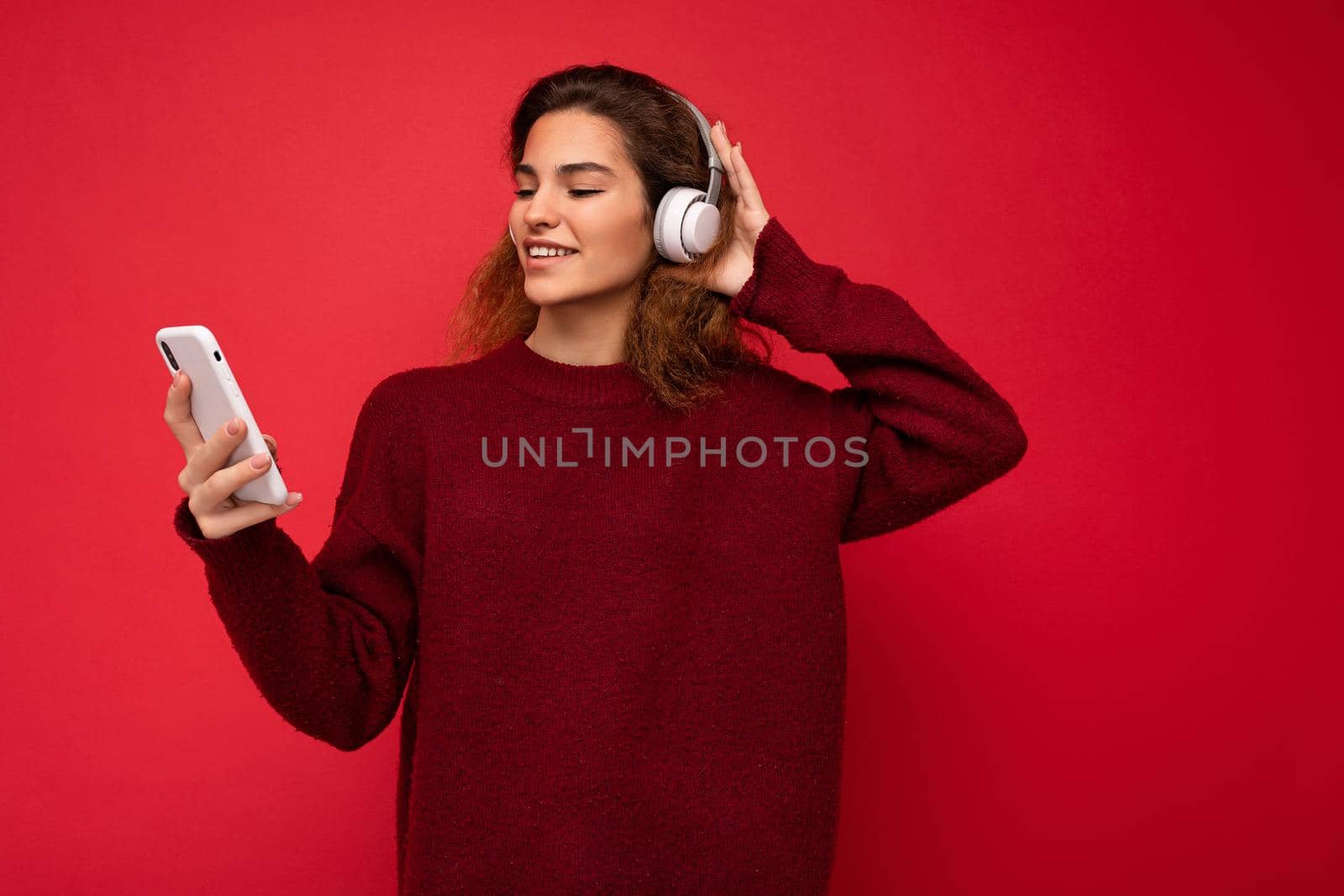 Photo of beautiful joyful smiling young woman wearing stylish casual clothes isolated over background wall holding and using mobile phone wearing white bluetooth headphones listening to music and having fun looking to the side.