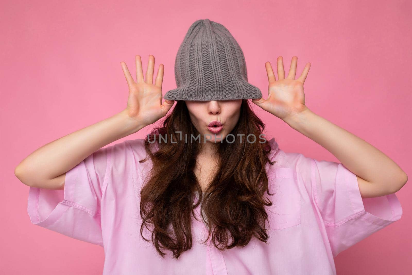 Photo of beautiful emotional young brunette woman standing isolated over pink background wall wearing pink shirt and gray hat and having fun.