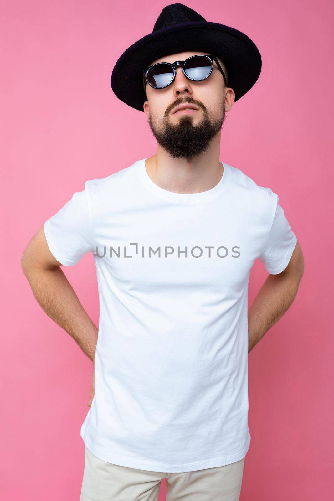 Vertical photo shot of handsome self-confident brunette unshaven young man with beard wearing casual white t-shirt for mockup black hat and stylish sunglasses isolated over pink background wall looking at camera by TRMK