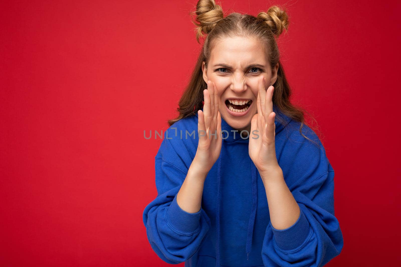 Photo of emotional angry young beautiful nice blonde female person with two horns with sincere emotions wearing trendy bright blue hoodie isolated over red background with empty space and yelling.