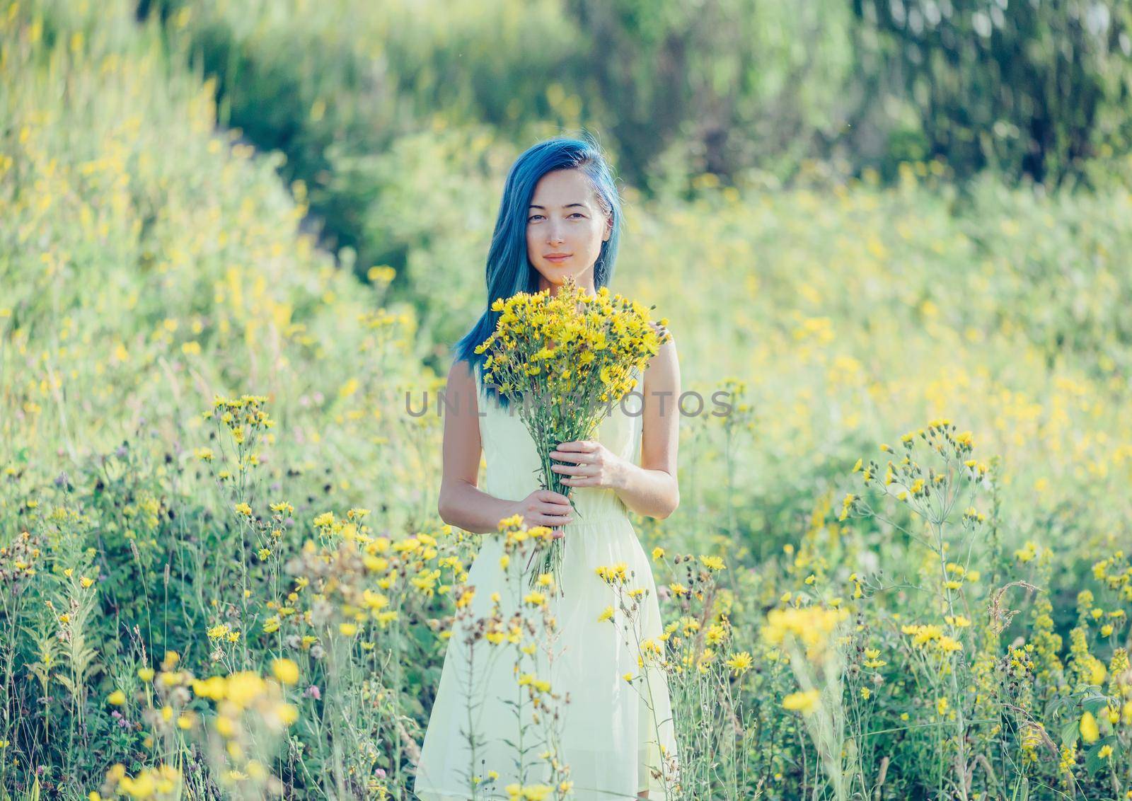 Smiling young woman with blue hair and a bouquet of yellow flowers standing in a field