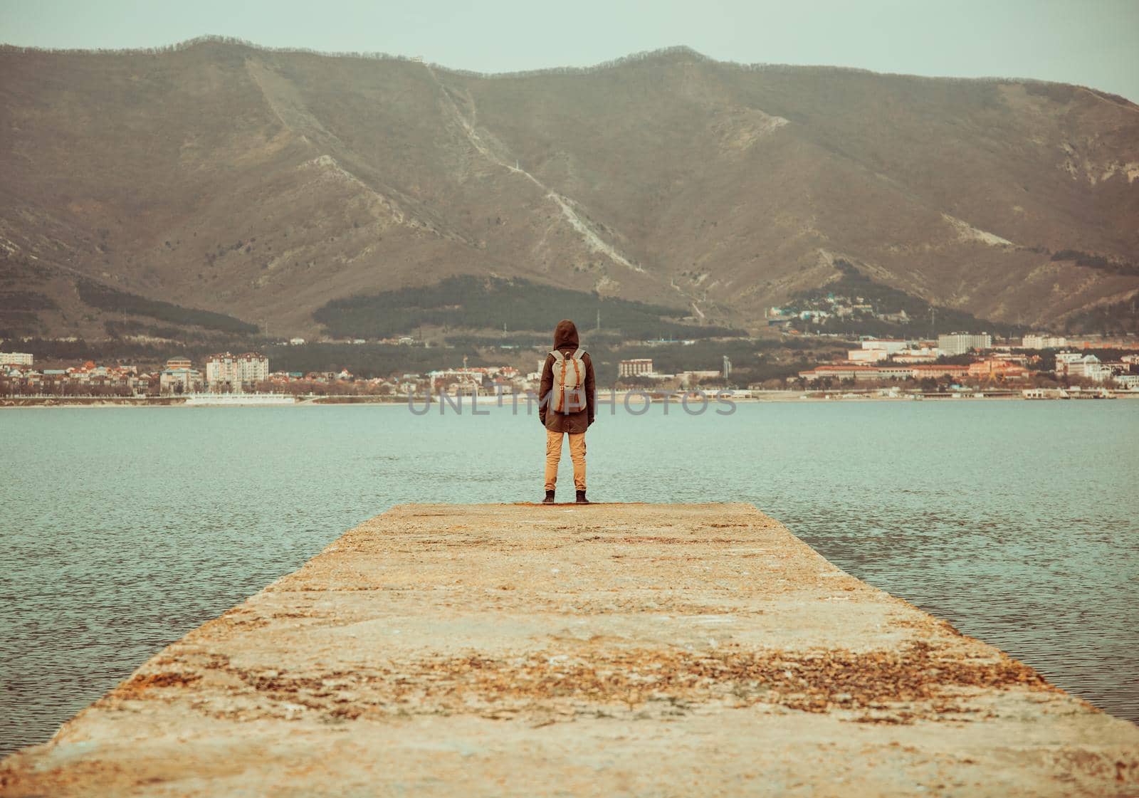 Unrecognizable traveler with backpack standing on pier at bay in front of mountains, rear view