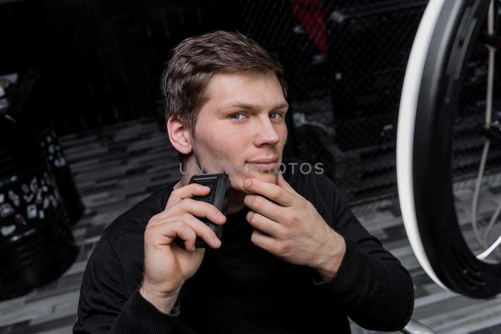 A serious young guy of Caucasian appearance with dark hair shaves with an automatic razor next to the ring lighting. Care for your appearance.