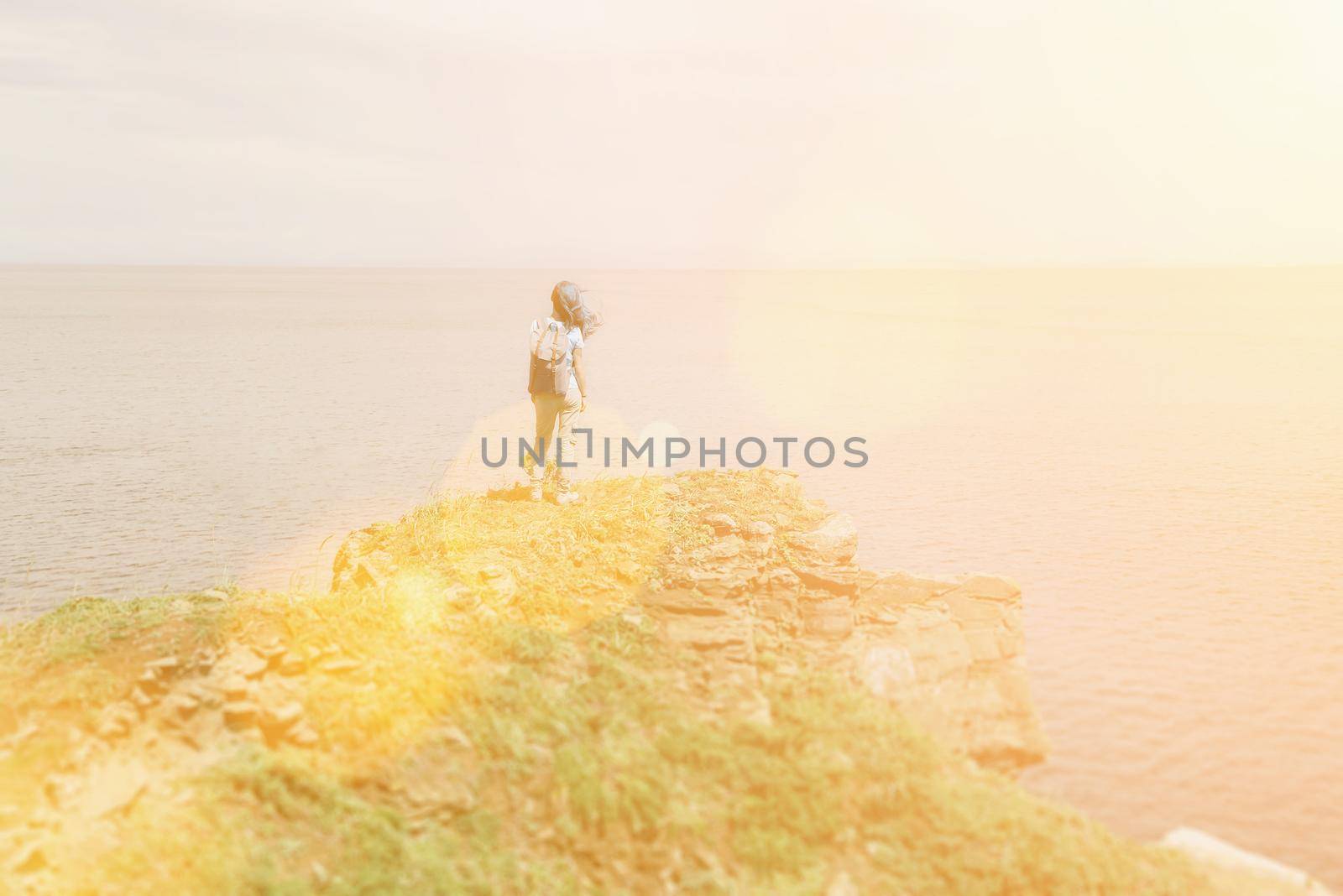 Hiker young woman with backpack standing on peak of mountain and enjoying view of sea in summer. Image with sunlight effect