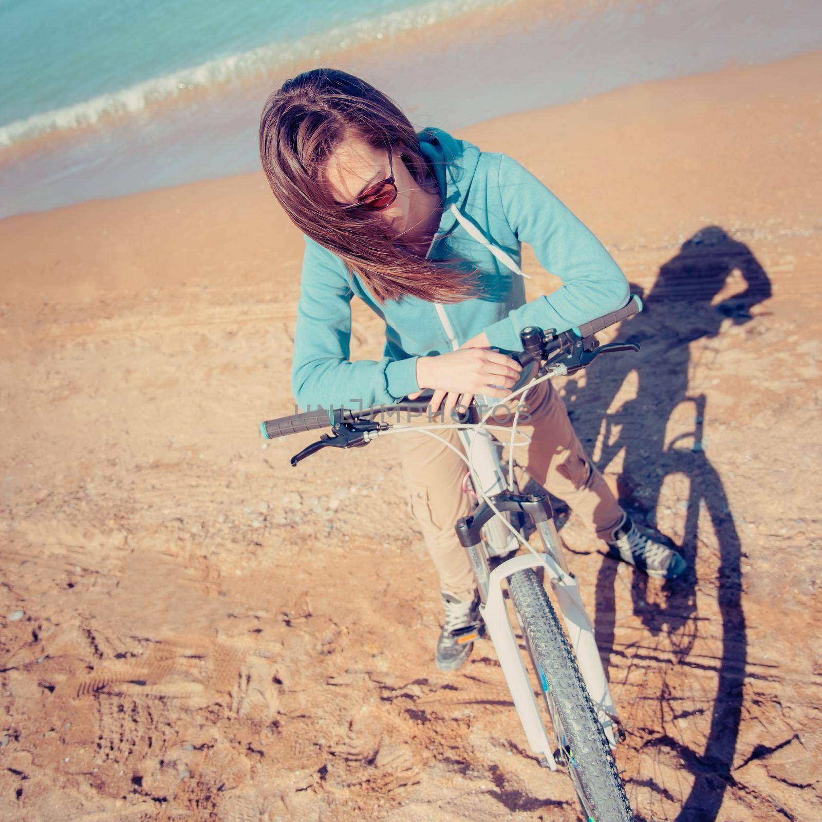 Young woman in sunglasses with bicycle standing on sand beach near the sea in summer