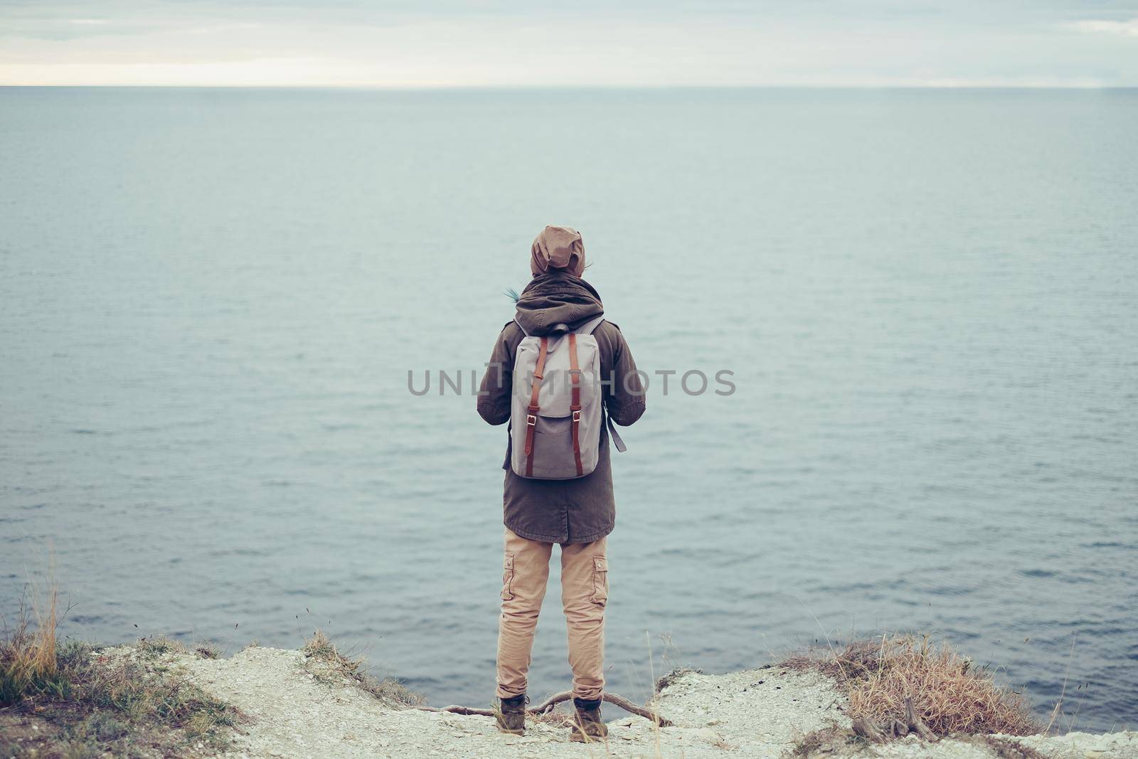 Unrecognizable traveler young woman with backpack standing on steep coast and enjoying view of sea, rear view. Traveler wearing in warm clothing
