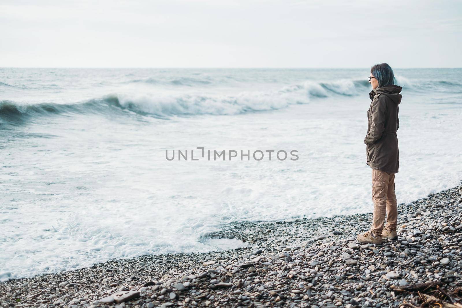 Traveler young woman standing on pebble coast and looking at sea waves in windy weather