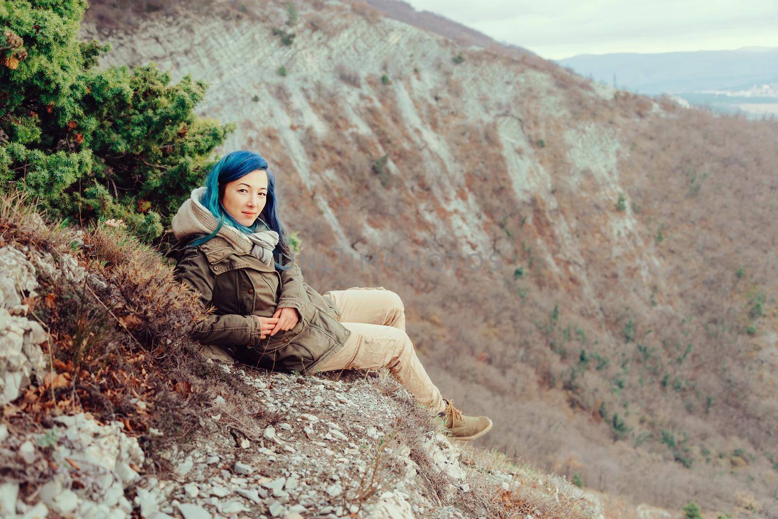 Smiling young woman wearing parka sitting in the mountains and looking at camera. Space for text in right part of image