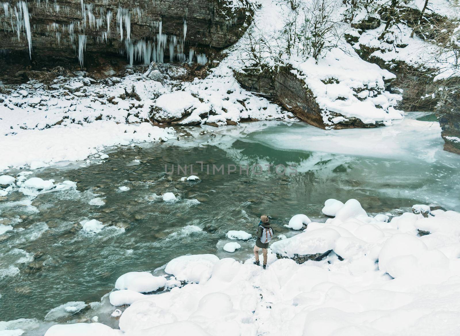 Hiker girl with backpack standing near the mountain river and enjoying view of nature in winter.