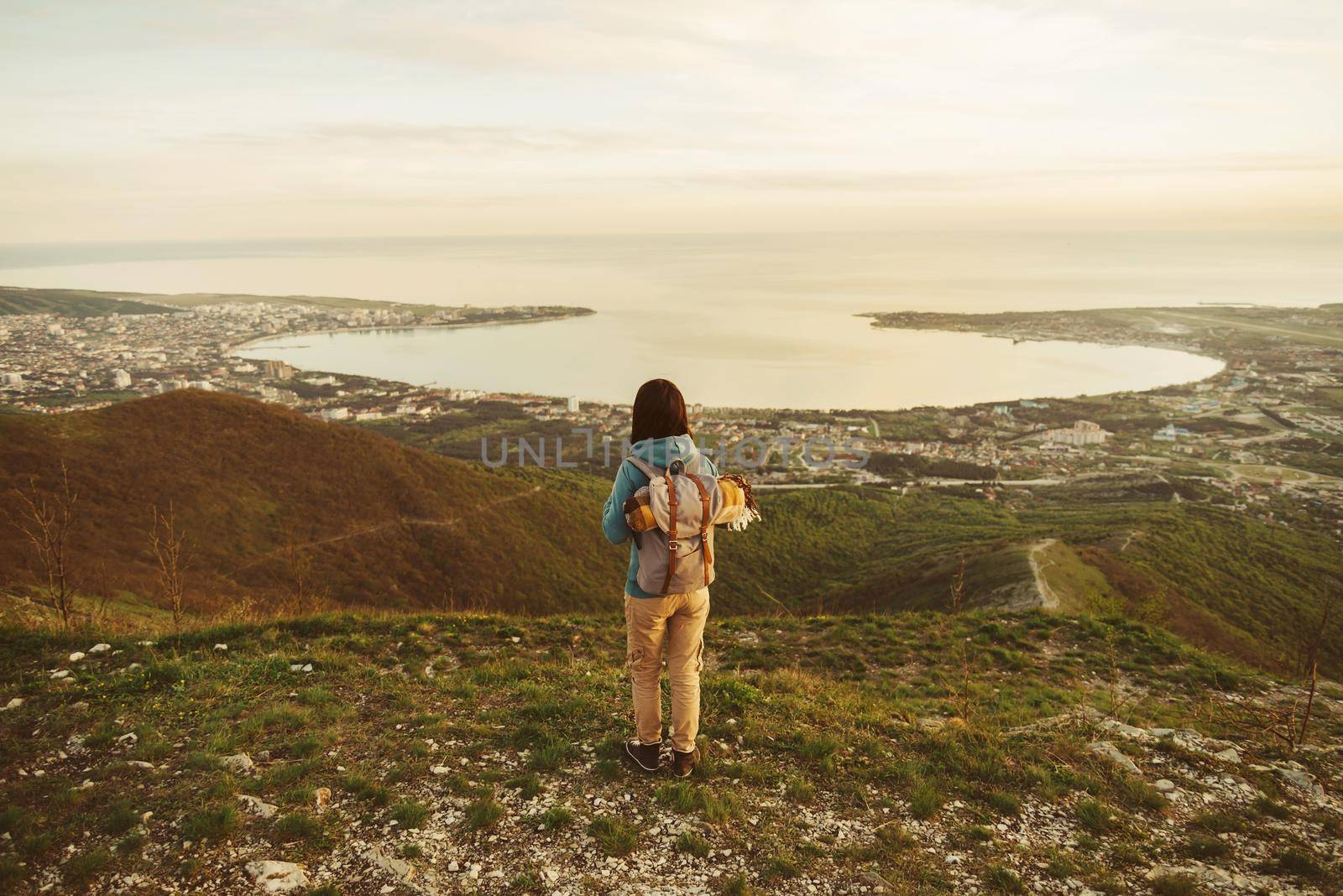 Traveler young woman with backpack standing on peak of mountain above sea bay and enjoying view of nature, rear view.