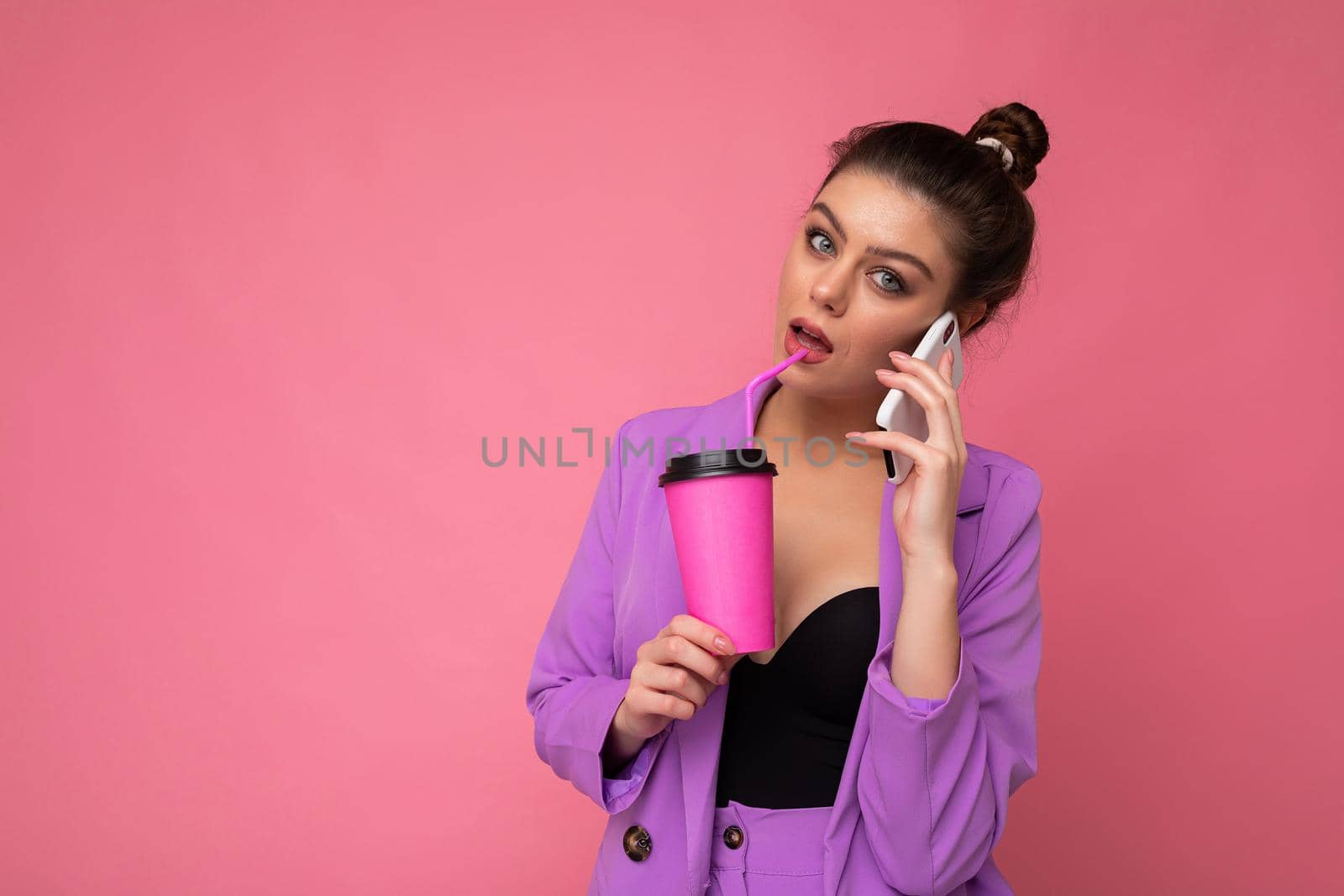 Portrait of pretty brunette woman wear trendy purple jacket, talking on smart phone, holding takeaway coffee in paper cup, isolated over pink background blank space for advertising.