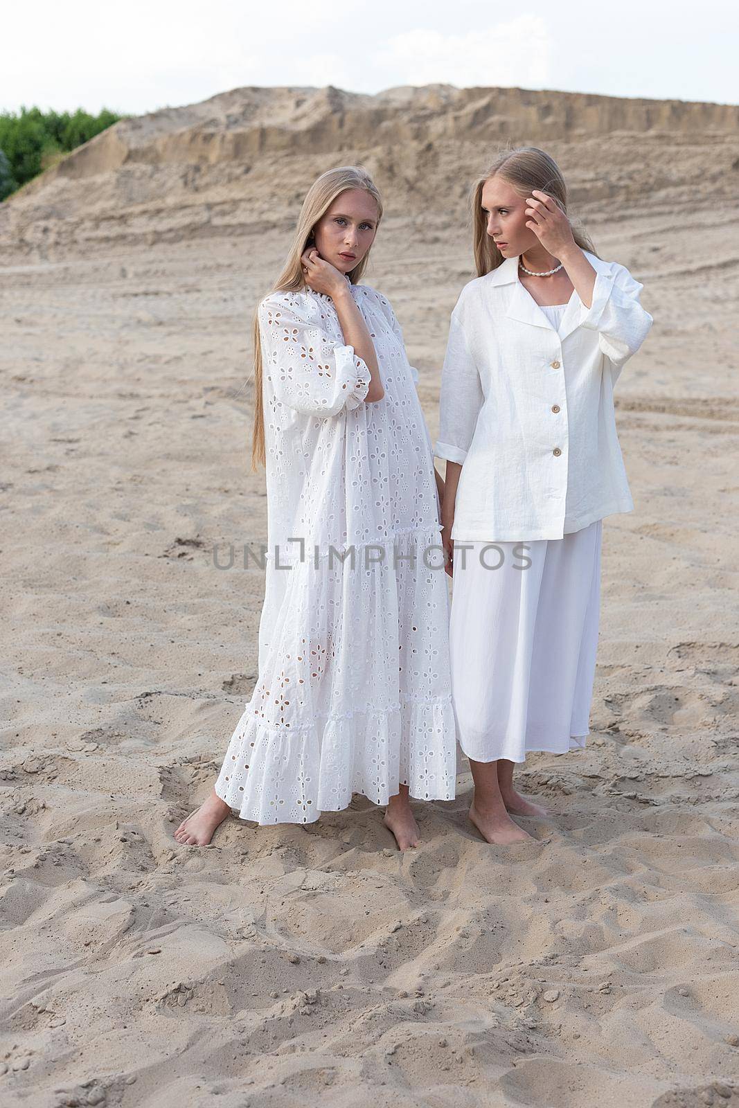 two young pretty twins with long blond hair posing at sand quarry in elegant white dress, skirt, jacket. stylish fashion photoshoot, summer photosession. identical sisters spend time together outdoors