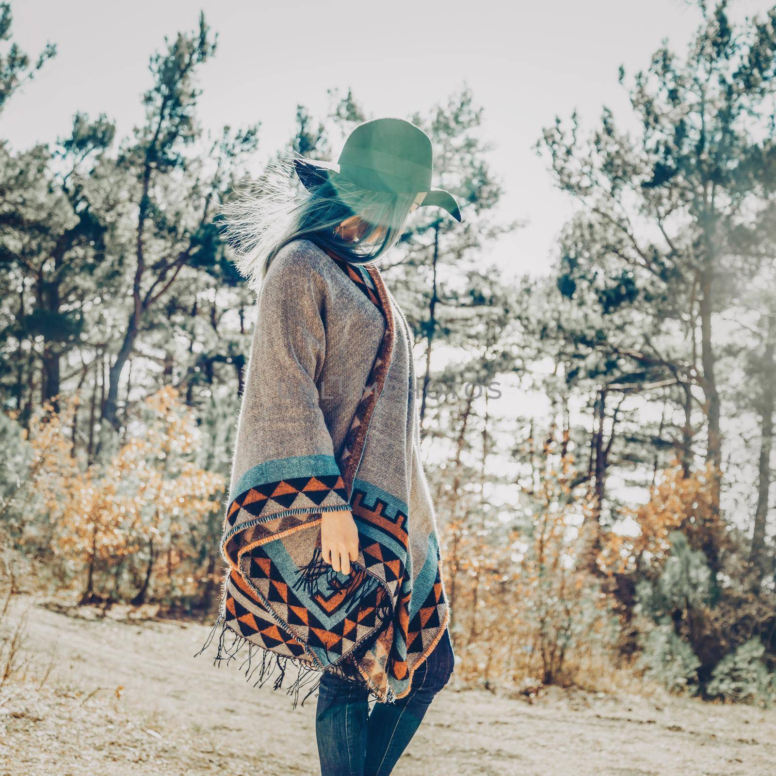 Fashionable young woman in hat and poncho walking in forest among pine trees