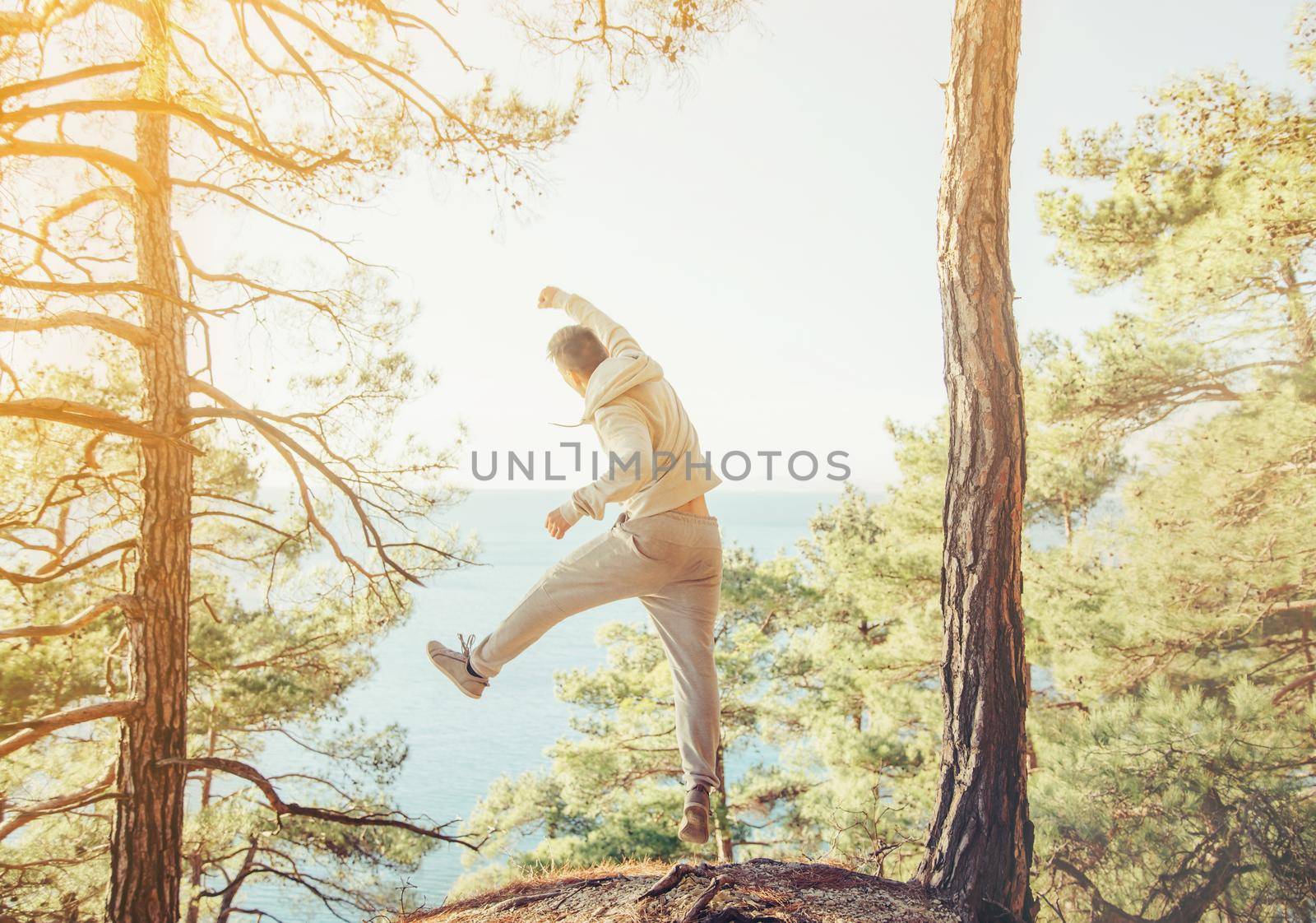 Happy sporty guy jumping on coast in the forest among pine trees in front of sea at sunny day, rear view