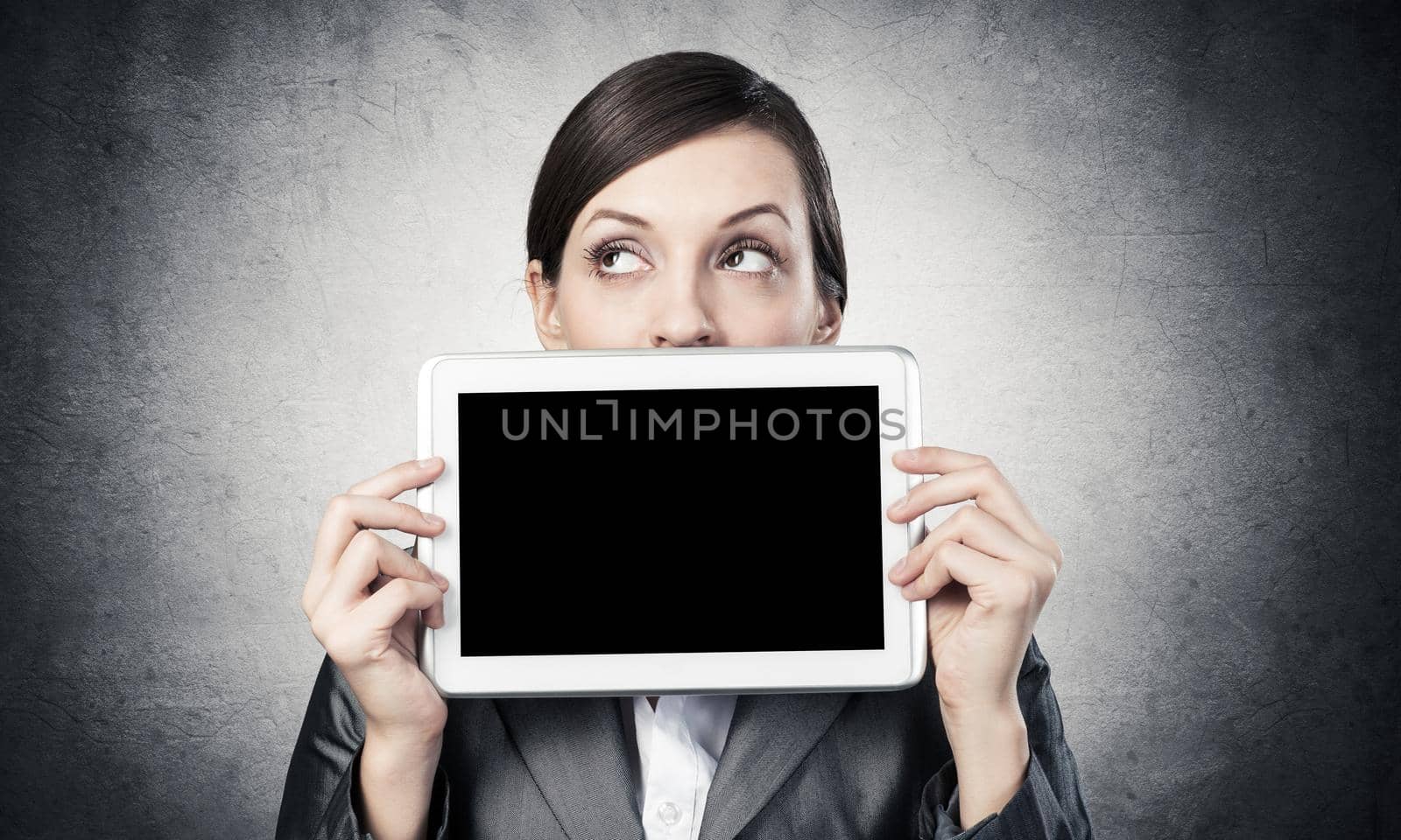 Businesswoman holding tablet computer with blank screen. Beautiful woman in business suit show tablet PC near her face. Corporate businessperson on grey wall background. Digital technology layout.