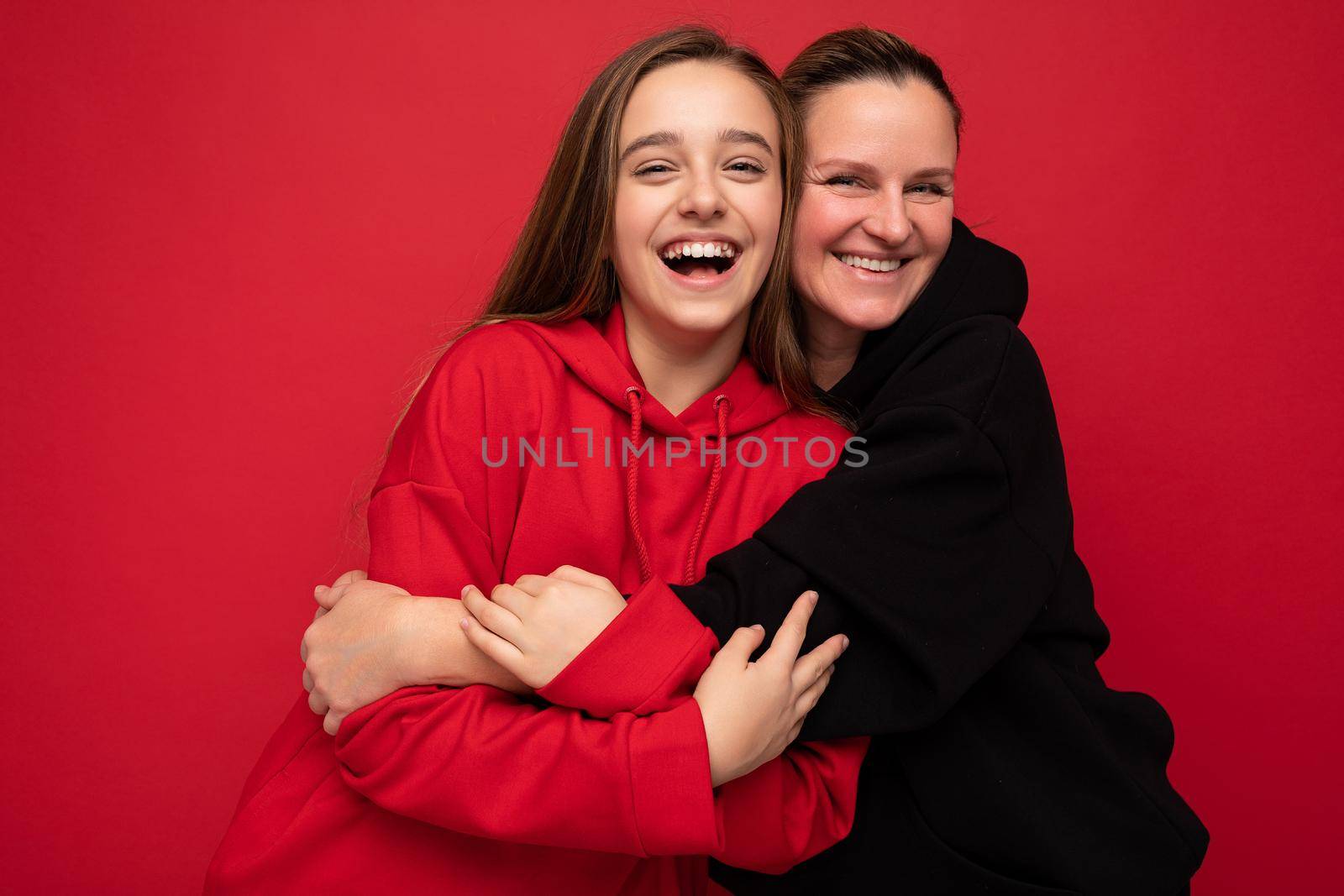 Photo shot of pretty positive smiling brunette female teenager wearing stylish red hoodie and adult brunette woman wearing trendy black hoodie standing isolated over red background wall looking at camera and having fun by TRMK