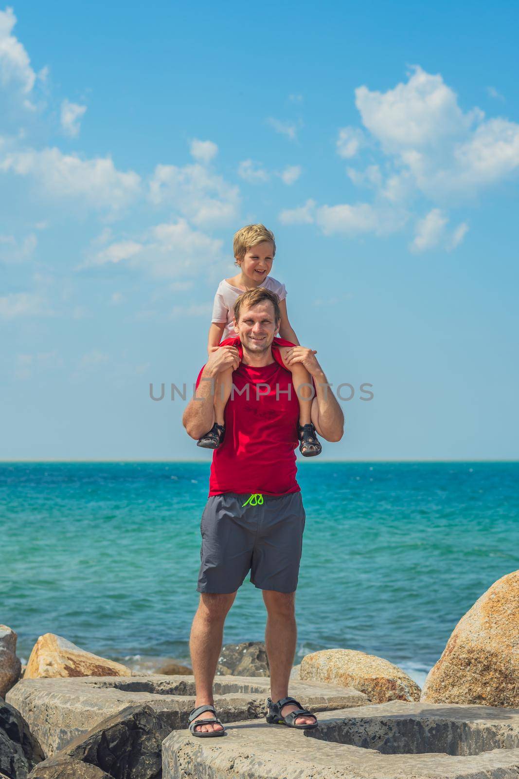 Happy smile father hugs holds on shoulders boy male child stand blue lazur sea panorama skyline horizon sunshine day clear sky. Travel, relationship, dad responsibilities, influence on son worldview.